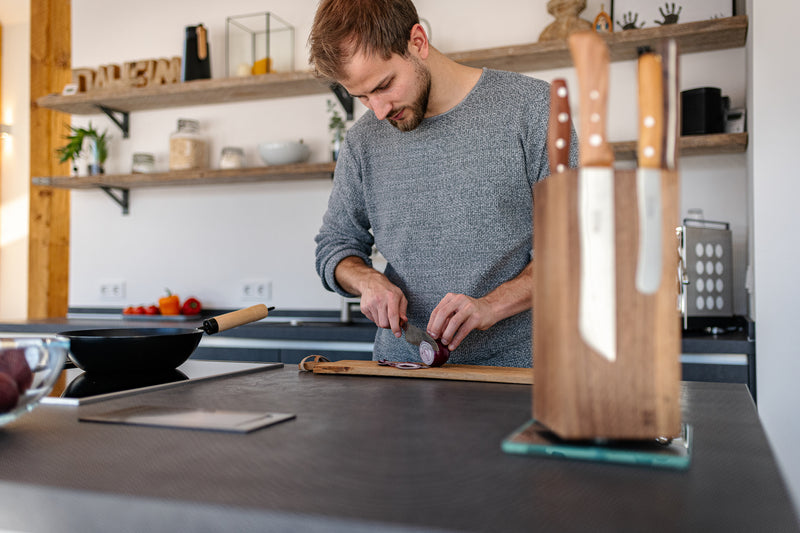 Ein Mann in einer Küche der eine Zwiebel schneidet. Im Vordergrund ein Nussbaum Messerhalter mit Messer der unscharf fotografiert ist.